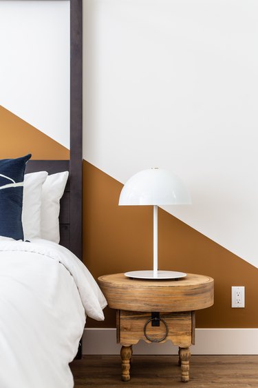 Modern rustic bedroom with dark wood headboard, rustic wood nightstand, white lamp, gold and white wall.